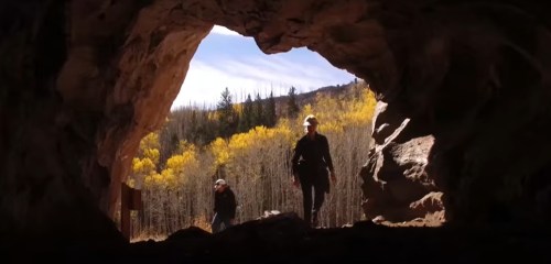 Two hikers exit a cave, surrounded by trees with vibrant autumn foliage in the background.