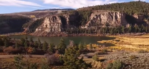 Scenic view of a lake surrounded by cliffs and trees, with mountains in the background under a clear sky.