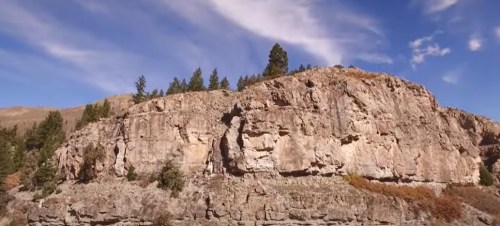 A rocky cliff with sparse trees under a blue sky with wispy clouds.