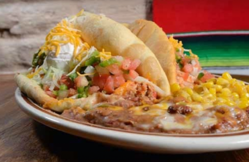 A plate of tacos topped with cheese, lettuce, and pico de gallo, served with refried beans and corn on the side.