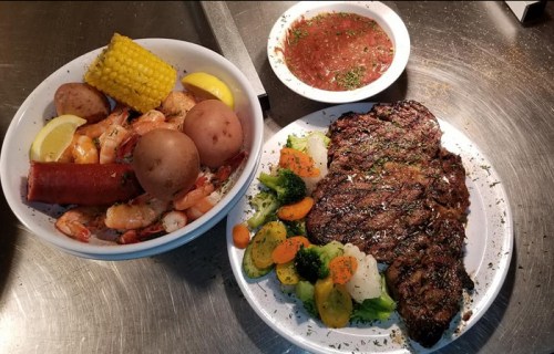 A plate of grilled steak with mixed vegetables, alongside a bowl of shrimp, potatoes, corn, and dipping sauce.