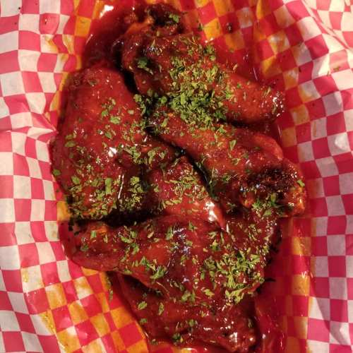 A basket of chicken wings coated in a glossy red sauce, garnished with green herbs, on a red and white checkered paper.