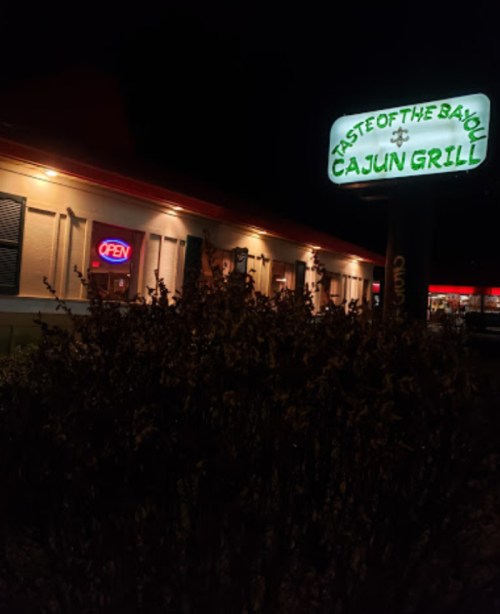 A nighttime view of "Taste of the Bayou Cajun Grill" with an illuminated "Open" sign and surrounding bushes.