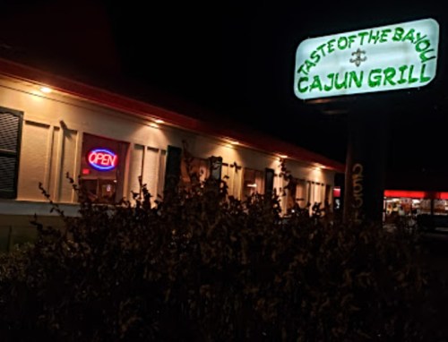 A nighttime view of "Taste of the Bayou Cajun Grill" with an illuminated "Open" sign and surrounding greenery.