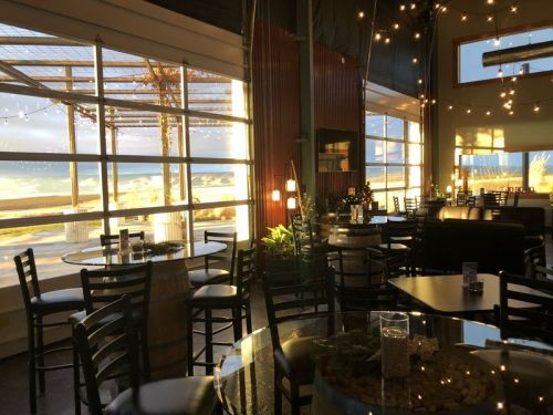 Cozy restaurant interior with tables, chairs, and large windows showcasing a beach view and string lights.