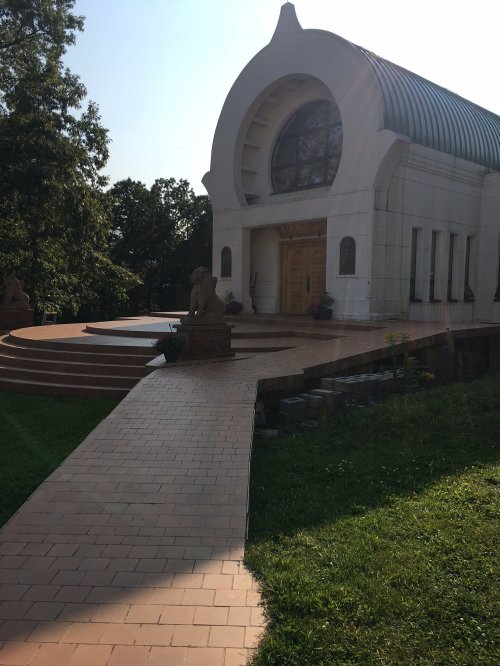 A modern building with a curved roof, large windows, and a stone pathway leading to its entrance, surrounded by greenery.
