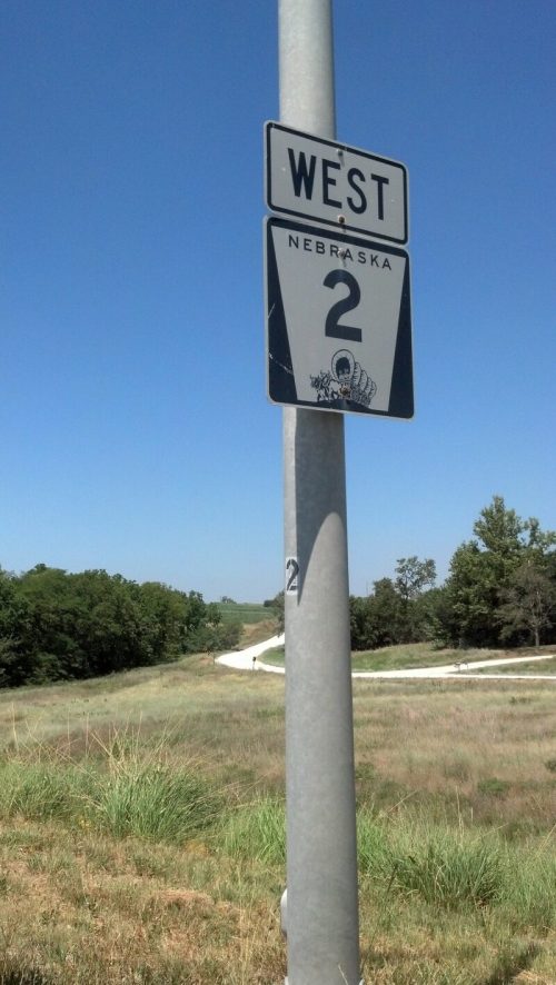 Sign for Nebraska Highway 2 West, mounted on a pole, with a clear blue sky and green landscape in the background.