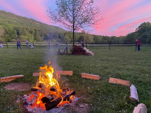 A campfire burns in a grassy area, with people enjoying the sunset and mountains in the background.