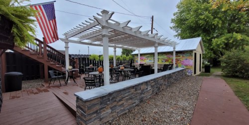 Outdoor seating area with a pergola, stone wall, and an American flag, surrounded by greenery and a mural.