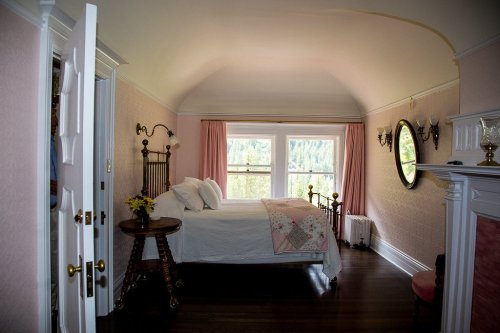 Cozy bedroom with a bed, pink walls, large windows, and a vintage mirror, featuring a warm, inviting atmosphere.