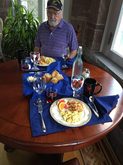 An elderly man sits at a dining table set with plates of food, drinks, and decorative items, enjoying a meal.