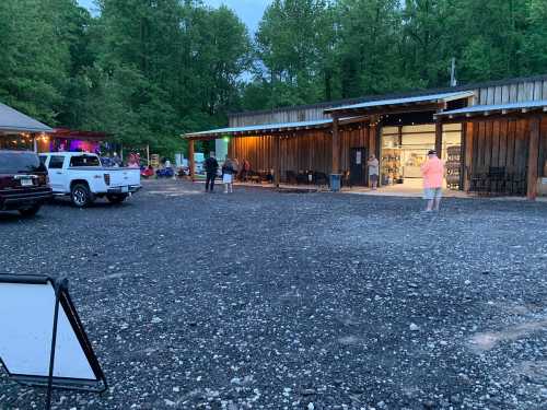 A rustic building with outdoor seating, surrounded by trees, and a gravel parking area with people and vehicles.