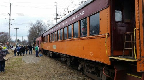 A vintage orange train car parked on a gravel path, with people gathered nearby on a cloudy day.