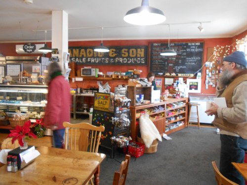 A cozy market interior with wooden tables, shelves of products, and a chalkboard menu. Holiday decorations are visible.