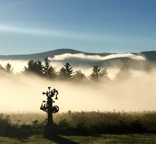 A misty landscape with a silhouette of a sculpture in the foreground and mountains in the background under a clear blue sky.