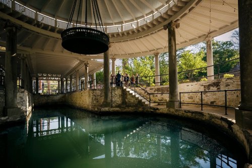 A circular, open-air structure with a pool, surrounded by columns and greenery, featuring visitors on a staircase.