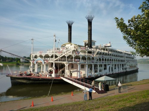 A large steamboat with two smokestacks docked by the river, featuring decorative bunting and a walkway for passengers.