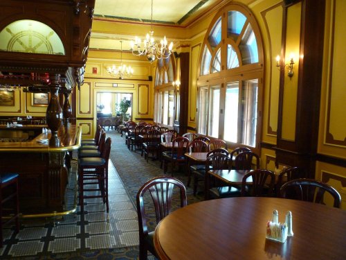 Elegant restaurant interior with wooden furniture, chandeliers, and large windows, creating a warm, inviting atmosphere.