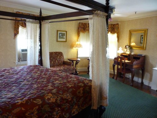 Cozy bedroom with a four-poster bed, armchair, desk, and warm lighting, featuring patterned decor and soft curtains.