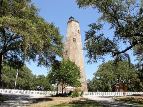 A tall lighthouse surrounded by trees and greenery under a clear blue sky. A white picket fence is visible in the foreground.