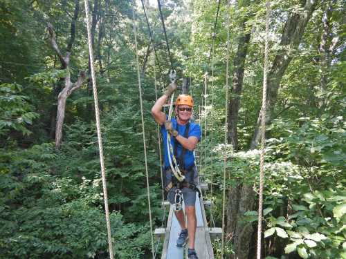 A smiling man in a helmet and harness stands on a zip line platform surrounded by lush green trees.