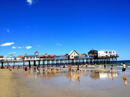 A sunny beach scene with a pier, people enjoying the water, and reflections on the wet sand under a clear blue sky.