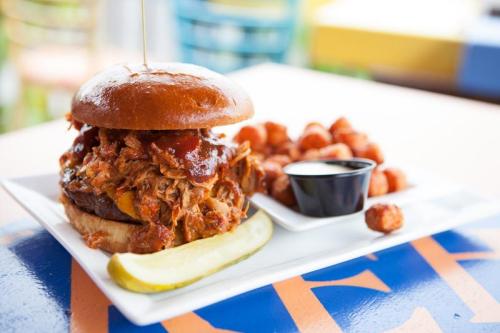 A pulled pork burger on a brioche bun, served with tater tots and a side of dipping sauce, garnished with a pickle.