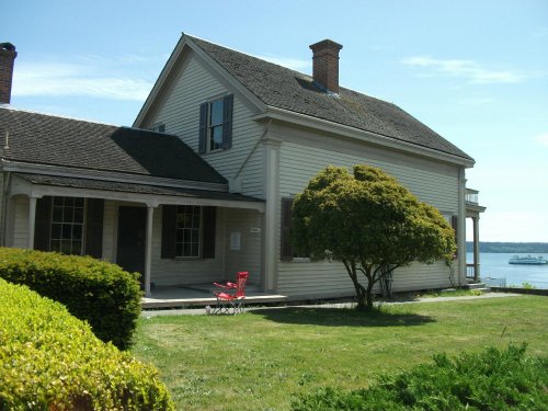 A historic house with a porch, surrounded by greenery, overlooking a body of water on a sunny day.
