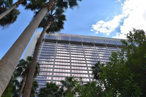 A tall building with reflective windows, surrounded by palm trees and blue sky with scattered clouds.