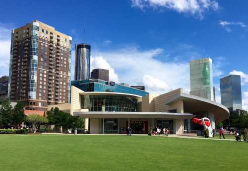 A modern building with a curved roof, surrounded by green grass and tall city skyscrapers under a blue sky.