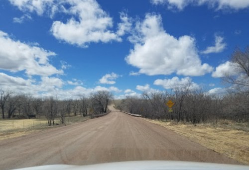 A dirt road stretches ahead under a blue sky with fluffy clouds, flanked by bare trees and warning signs.