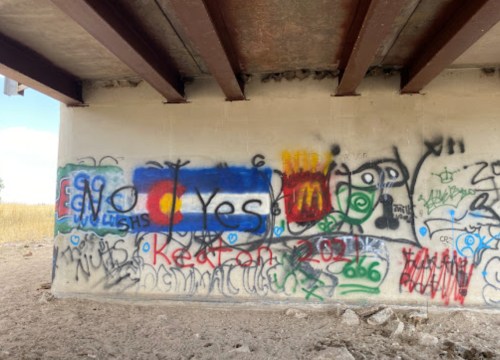 Colorful graffiti on a concrete wall, featuring the Colorado flag and various tags and messages.