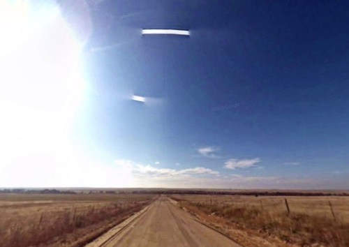 A wide dirt road stretches into the distance under a bright blue sky with a shining sun and scattered clouds.