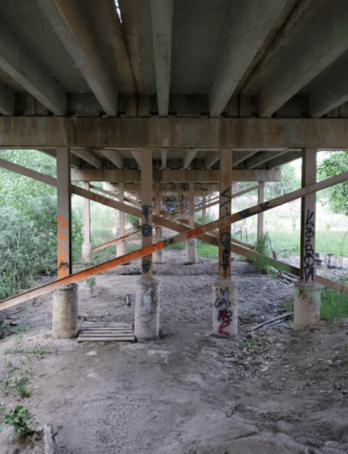 View of a concrete underpass with graffiti on pillars and wooden planks on the ground, surrounded by greenery.