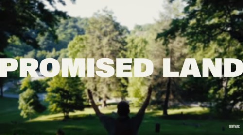A person stands with arms raised in a lush, green landscape, with the text "PROMISED LAND" prominently displayed.