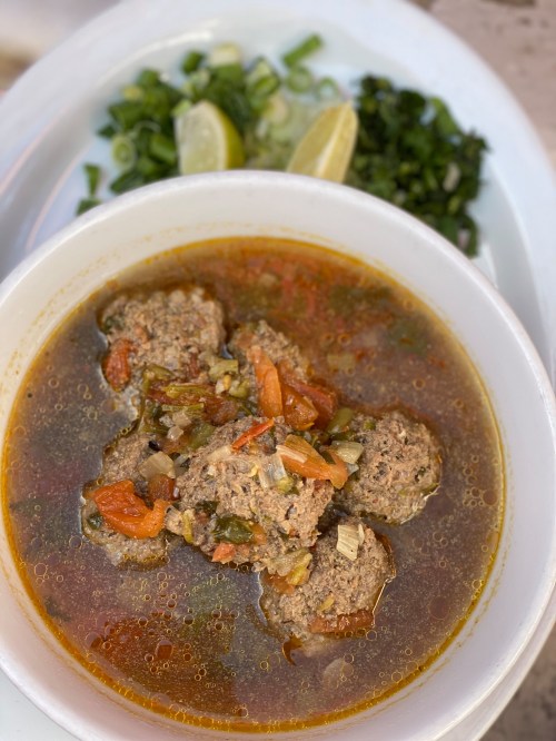 A bowl of meatball soup with vegetables, served with lime wedges and chopped green onions on the side.