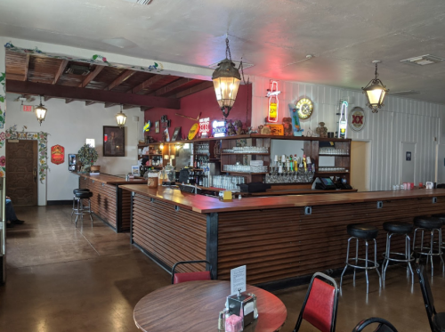 A cozy restaurant interior featuring a wooden bar, colorful decor, and vintage signs, with tables and chairs in view.