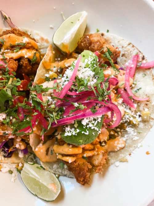 Two tacos filled with fried fish, topped with sauce, pickled onions, cilantro, and lime wedges on a white plate.
