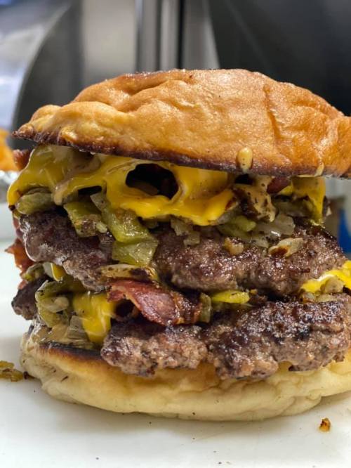 A towering burger with three beef patties, melted cheese, bacon, and pickles on a toasted bun.