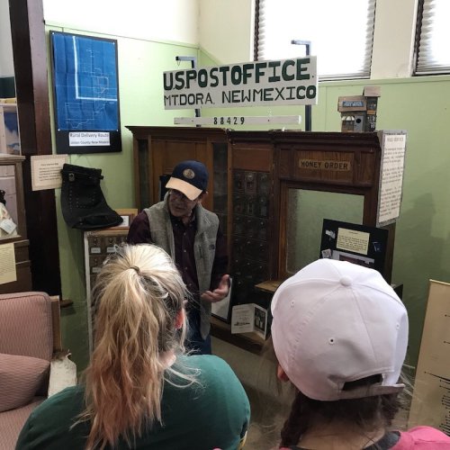 A man speaks to visitors in a small museum, showcasing historical items related to the Mt. Dora post office in New Mexico.