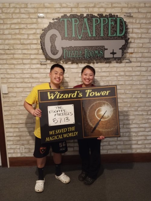 Two people smile while holding a sign that reads "Wizard's Tower" and celebrates their escape room achievement.