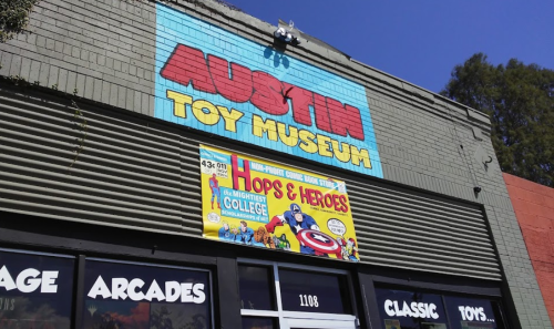 Colorful sign for the Austin Toy Museum, featuring "Hops & Heroes" event banner, with clear blue sky above.