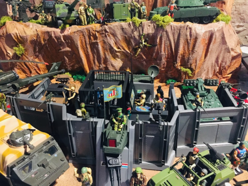 A detailed military diorama featuring soldiers, vehicles, and a fortified base set against a rocky backdrop.