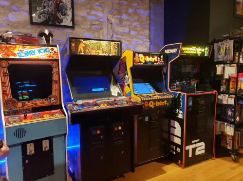A row of classic arcade machines, including Donkey Kong, Q*bert, and Terminator 2, set against a stone wall.