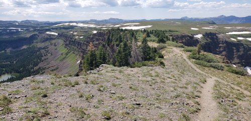 A scenic view from a rocky ridge, featuring green trees, distant mountains, and patches of snow under a cloudy sky.