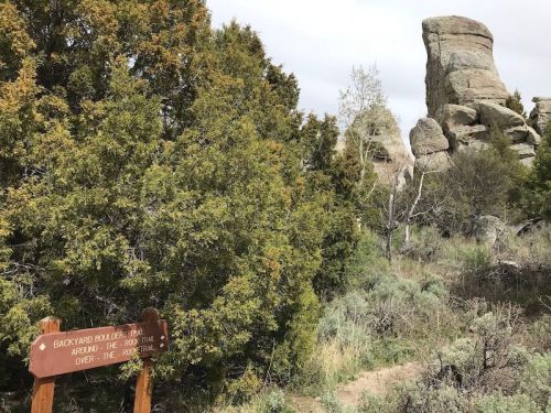 A wooden sign in a natural landscape, with rocky formations and greenery in the background under a cloudy sky.