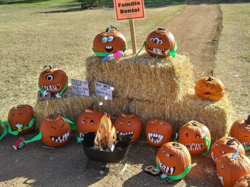 A display of carved pumpkins with teeth, set on hay bales, featuring a sign for "Familia Dental" and a small fire.