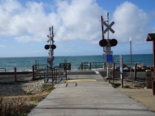 A pathway leads to the ocean, flanked by railroad crossing signals and a clear blue sky with scattered clouds.