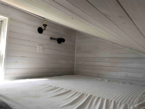 Cozy loft space with a bed, white walls, and a small window, featuring wall sconces for lighting.