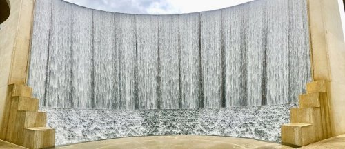 A large, curved waterfall cascades down a smooth stone wall, creating a serene and tranquil atmosphere.
